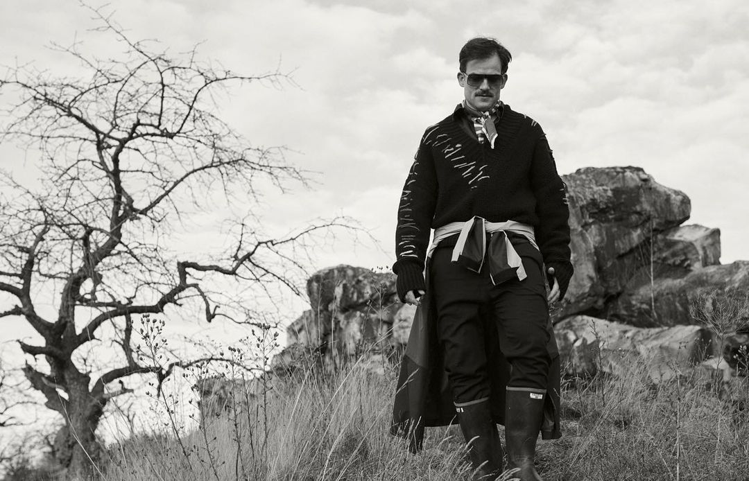 Man wearing sunglasses and a patterned sweater walking outdoors near rocky terrain and a bare tree