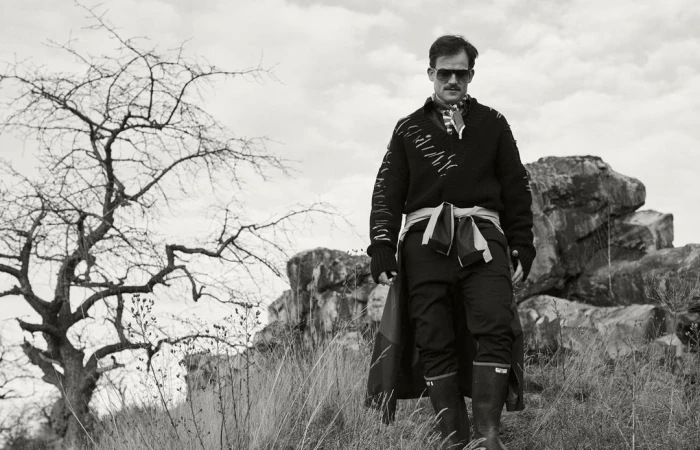 Man wearing sunglasses and a patterned sweater walking outdoors near rocky terrain and a bare tree