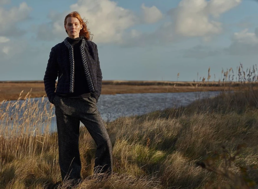 A woman with red hair stands in a field of tall grass near a body of water under a cloudy sky, wearing a dark tweed jacket and trousers.