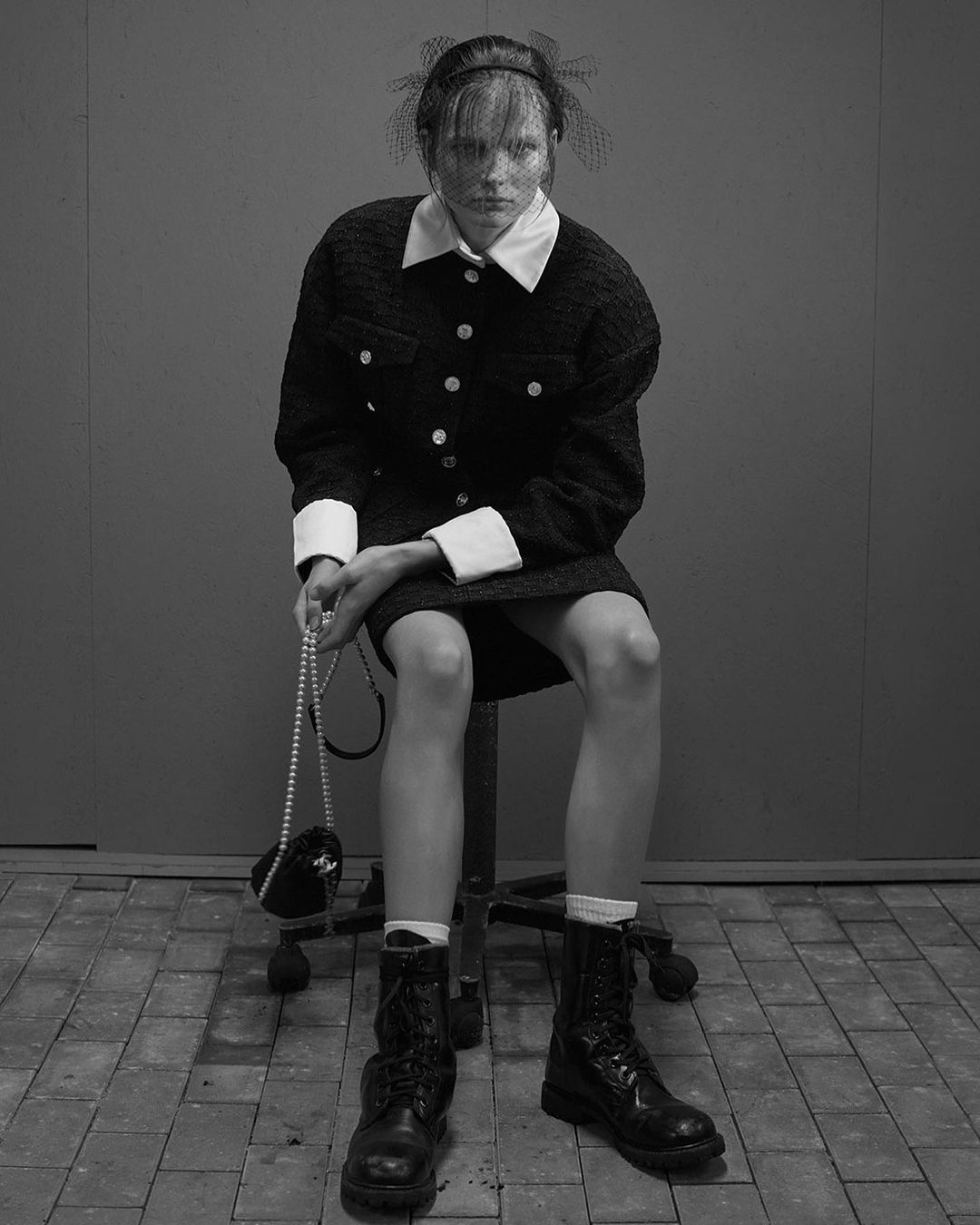 A fashionably dressed woman poses on a stool against a neutral backdrop in a studio, wearing a skirt suit, pearls, and combat boots.