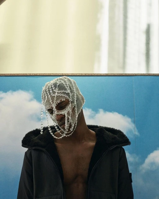 Close-up studio shot of a Black man wearing an ornate, beaded headpiece and a black jacket, set against a blue sky with clouds.