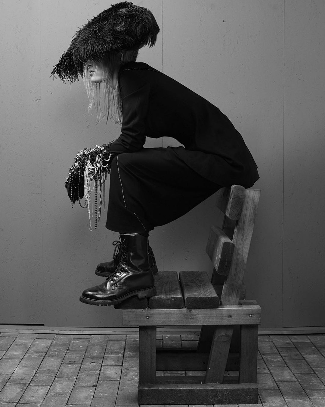 Black and white fashion shot of a model seated on a wooden chair wearing a feathered hat, dark clothing, and draped with strands of beads.