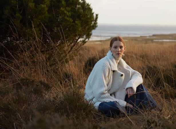 Red-haired model in a thick white knit sweater seated in a coastal field of dry grass by Ralph Mecke