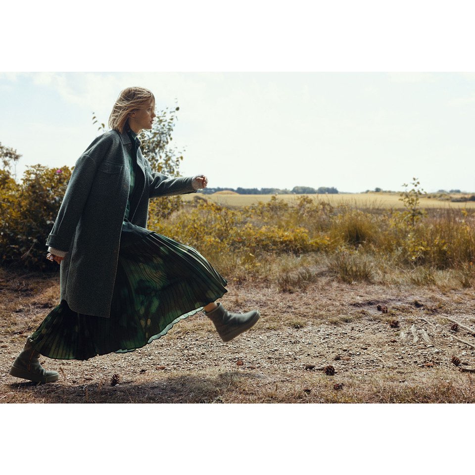 A blonde woman in a teal coat and green dress skips along a dirt path in a bright field surrounded by trees and tall grass.