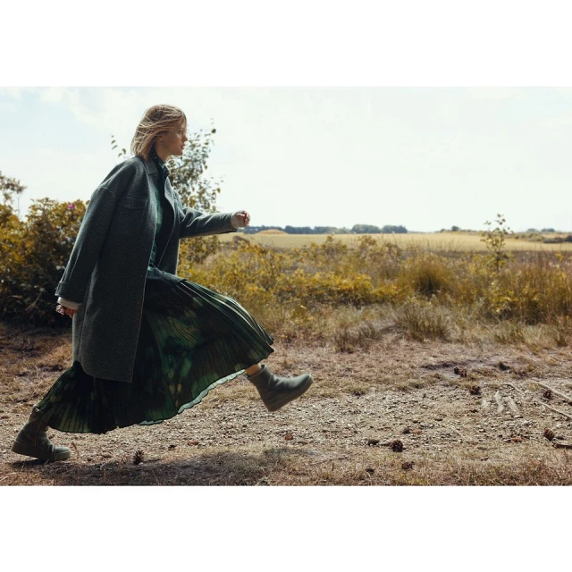 A blonde woman in a teal coat and green dress skips along a dirt path in a bright field surrounded by trees and tall grass.