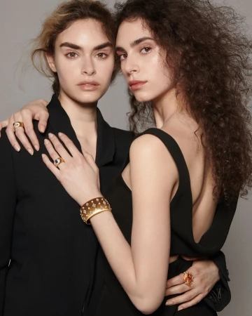 Two women pose in a studio, wearing black tops and gold jewelry, with one woman's arm around the other. The background is grey.