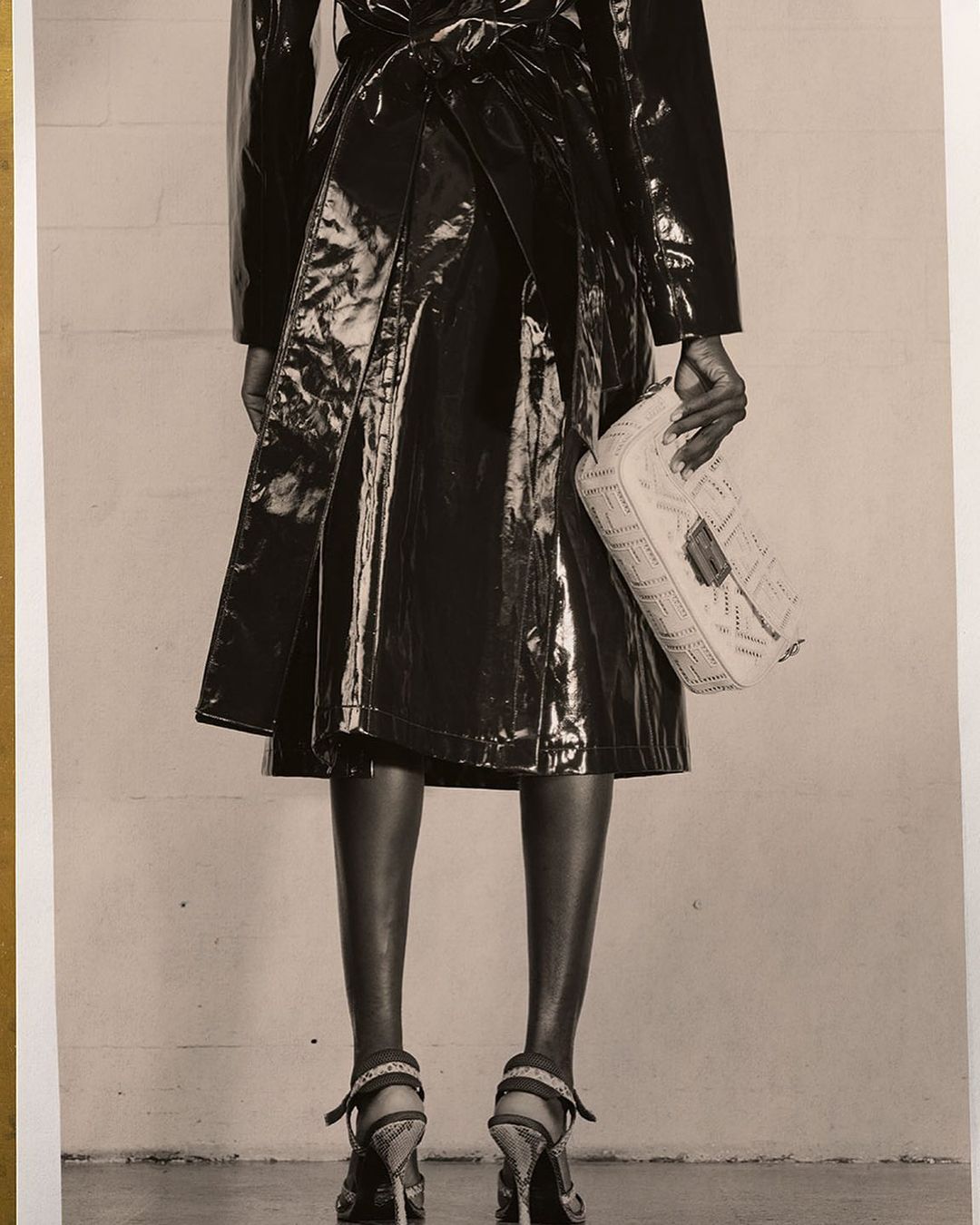 Back view of an adult woman wearing a shiny trench coat, holding a purse, and posing in high heels against a neutral background.