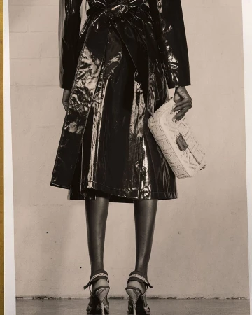 Back view of an adult woman wearing a shiny trench coat, holding a purse, and posing in high heels against a neutral background.