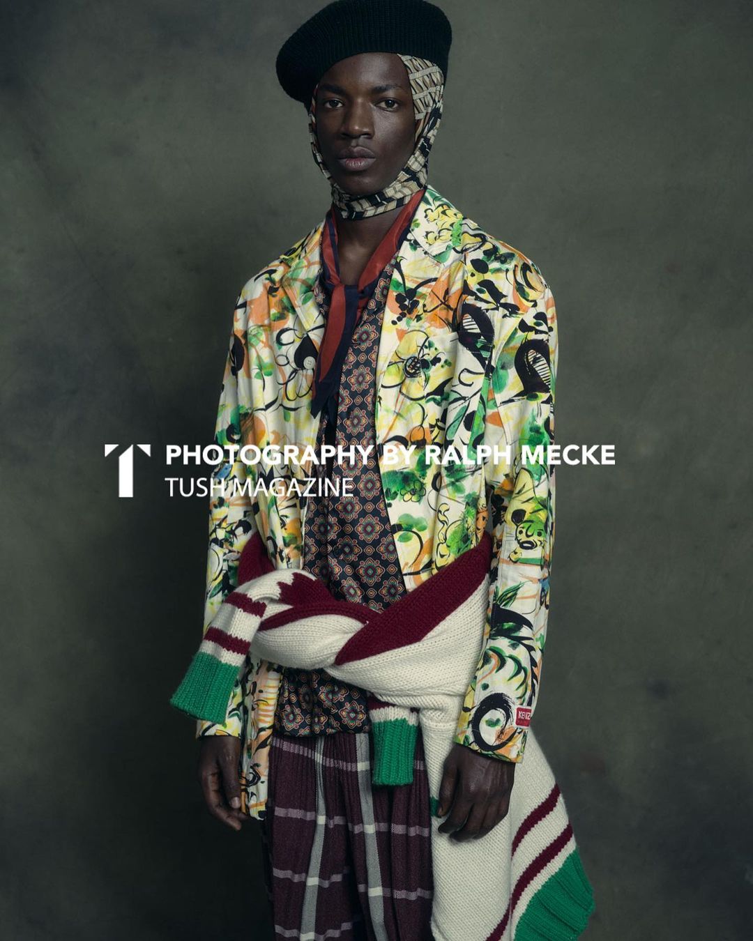 Black man wearing a patterned floral jacket, beret, and a sweater tied around his waist against a textured dark background.