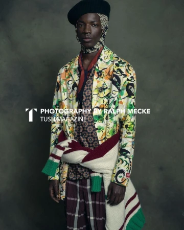 Black man wearing a patterned floral jacket, beret, and a sweater tied around his waist against a textured dark background.