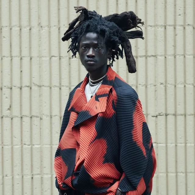 Portrait of an adult male with dreadlocks wearing a graphic patterned jacket against a neutral industrial background, medium shot.