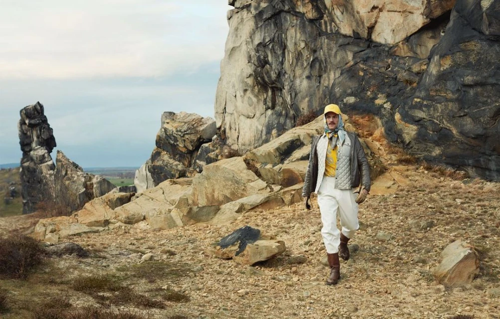 Man in vintage fashion walks on rocky terrain near large rock formations under a cloudy sky. Outdoors adventure and style.