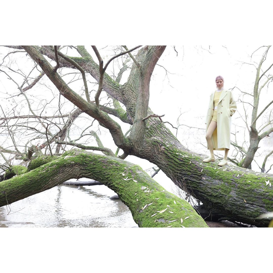 A fair-skinned adult female model wearing a yellow outfit and coat poses atop a mossy fallen tree in an outdoor water environment.