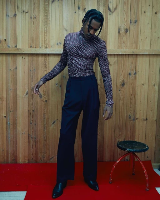 Adult male in patterned turtleneck and black dress pants in a modern fashion shot with a wooden background and a small stool.