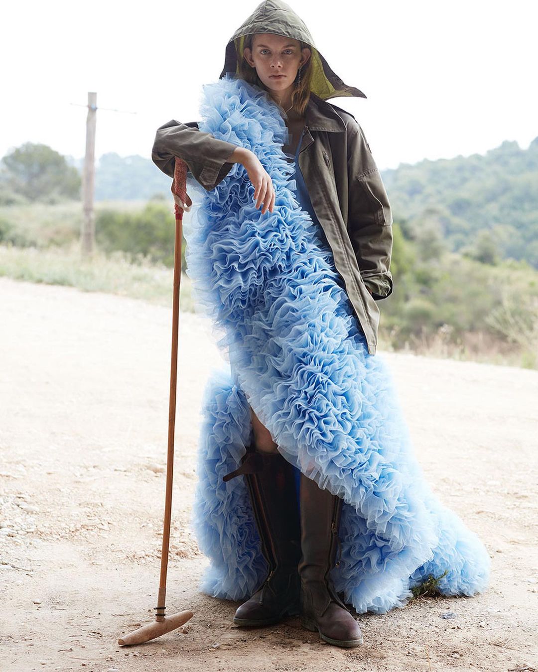 Fashionable woman in a blue ruffled dress with a hooded coat, standing outdoors with a rustic tool, creating a striking contrast.