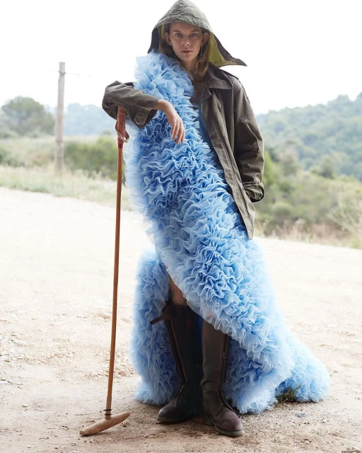 Fashionable woman in a blue ruffled dress with a hooded coat, standing outdoors with a rustic tool, creating a striking contrast.