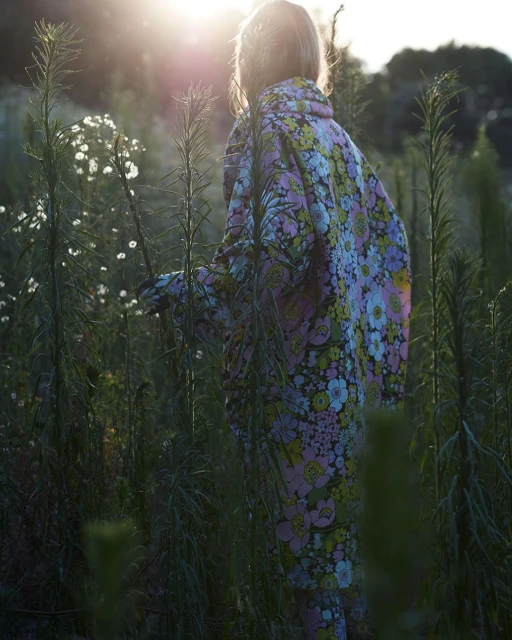 A young person in a colorful floral outfit stands in a field of tall green plants with white flowers, bathed in sunlight at sunset.