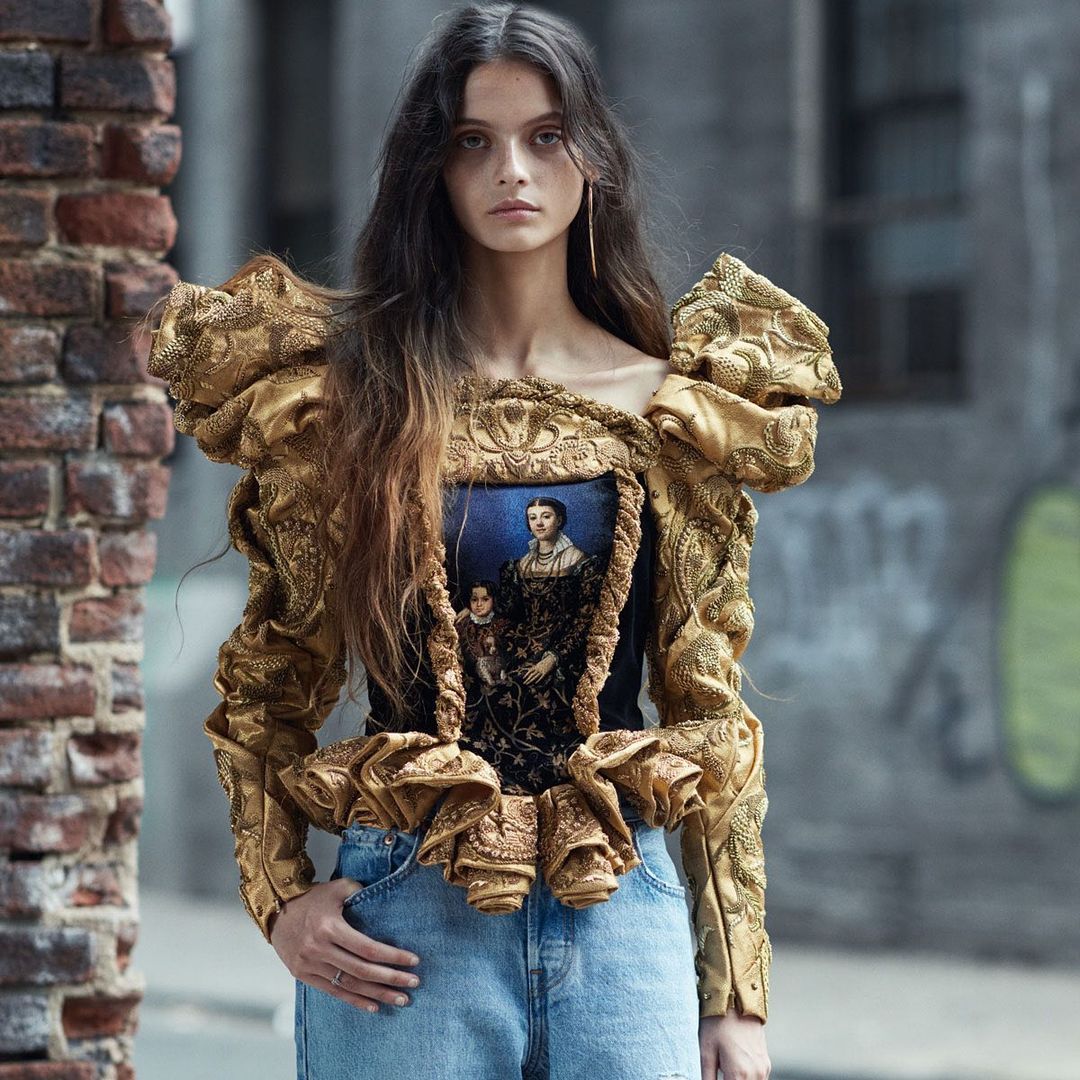 Brunette woman poses in an urban setting wearing a black top with a historical figure printed on it with gold ornate ruffles.