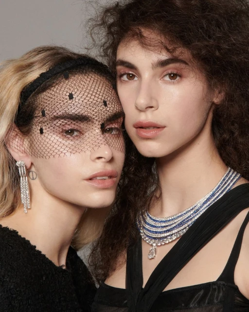 Two women with elegant makeup and jewelry pose indoors. One wears a black veil with dots and chandelier earrings; the other, a tiered necklace.