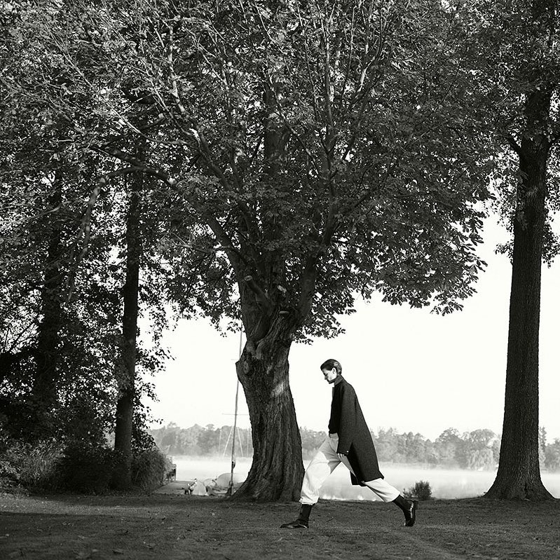 Model in dark coat and light trousers walking under a large leafy tree by a lake, photographed by Ralph Mecke in black and white.