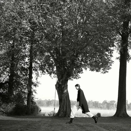 Model in dark coat and light trousers walking under a large leafy tree by a lake, photographed by Ralph Mecke in black and white.