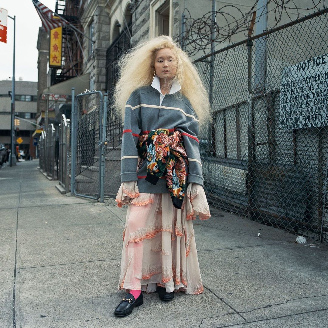 Fashionable Asian woman with bleached hair in a stylish skirt and grey sweater, posing on an urban sidewalk, wearing a floral jacket.