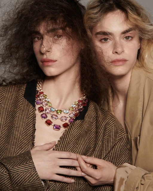 Two women wearing gemstone necklaces and jackets pose indoors, with neutral makeup and soft diffused lighting, highlighting fashion and beauty.