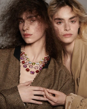 Two women wearing gemstone necklaces and jackets pose indoors, with neutral makeup and soft diffused lighting, highlighting fashion and beauty.