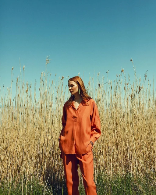 A woman with red hair in an orange linen suit stands in front of tall reeds against a blue sky. Fashion editorial portrait, outdoor setting.