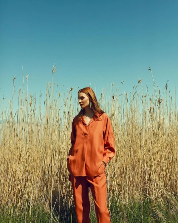 A woman with red hair in an orange linen suit stands in front of tall reeds against a blue sky. Fashion editorial portrait, outdoor setting.