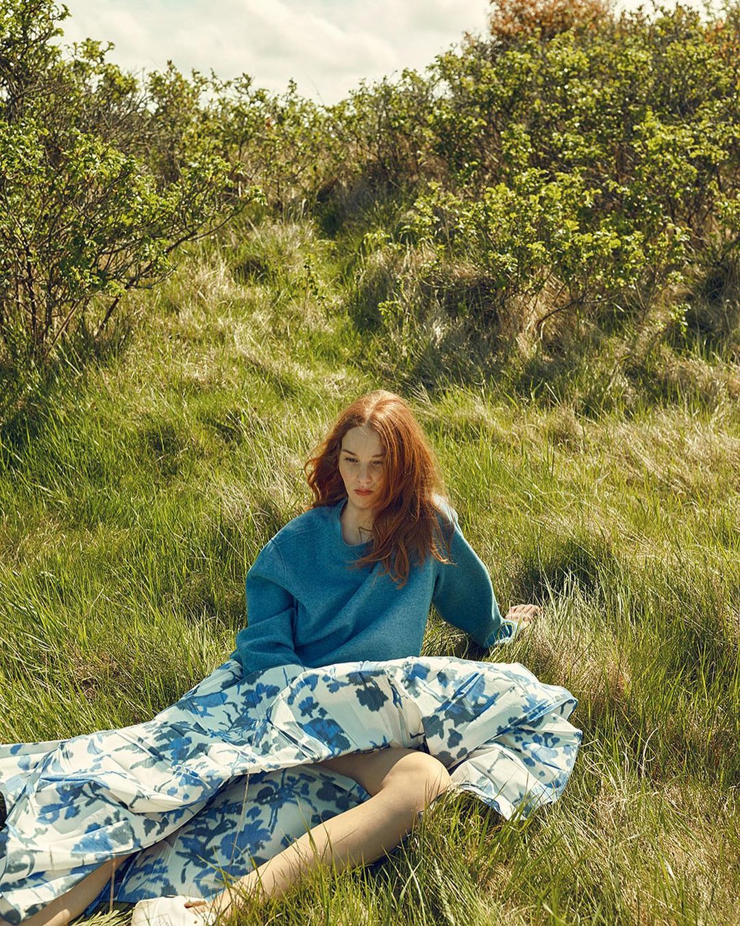 A red-haired woman in a blue sweater and floral skirt sits in a grassy field, enjoying the warm sunlight and natural scenery.
