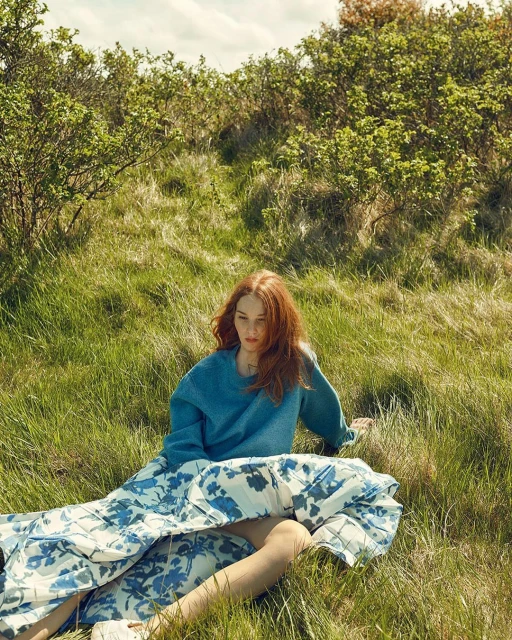 A red-haired woman in a blue sweater and floral skirt sits in a grassy field, enjoying the warm sunlight and natural scenery.