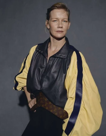 Portrait of a woman wearing a black and yellow leather jacket and monogram belt against a grey wall