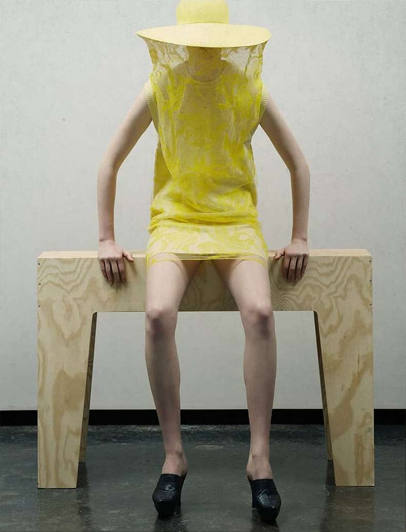 Person in a yellow hat and dress sits on a wooden bench in a studio with black shoes. Fashion shot with a textured yellow dress.