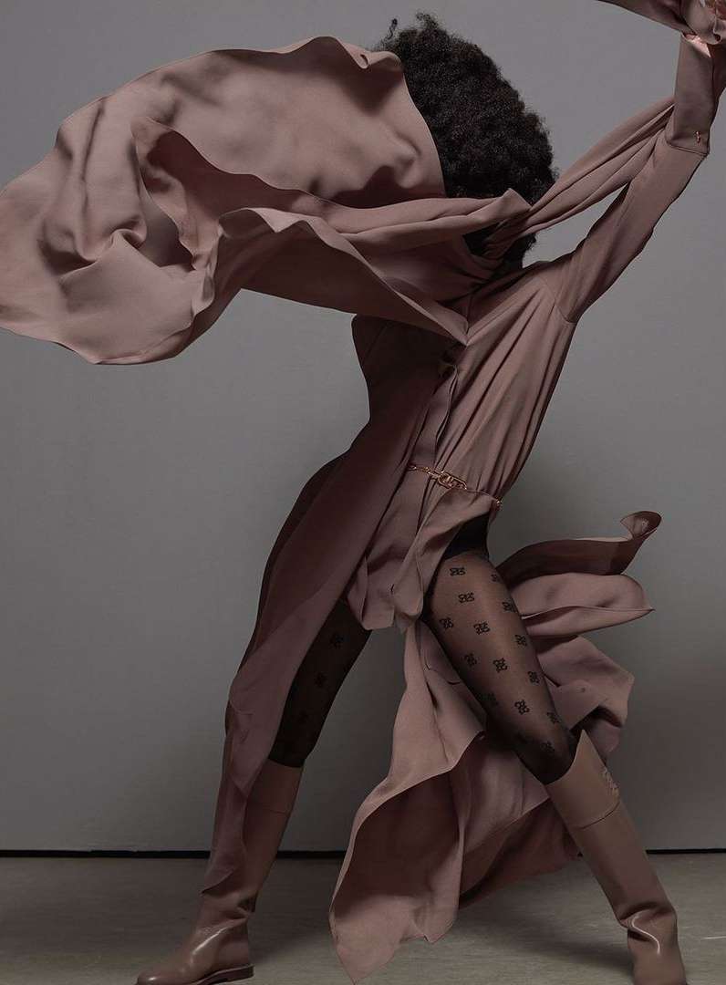 Fashionable person strikes dynamic pose in flowing mauve dress, black patterned tights, and matching boots against a grey backdrop.