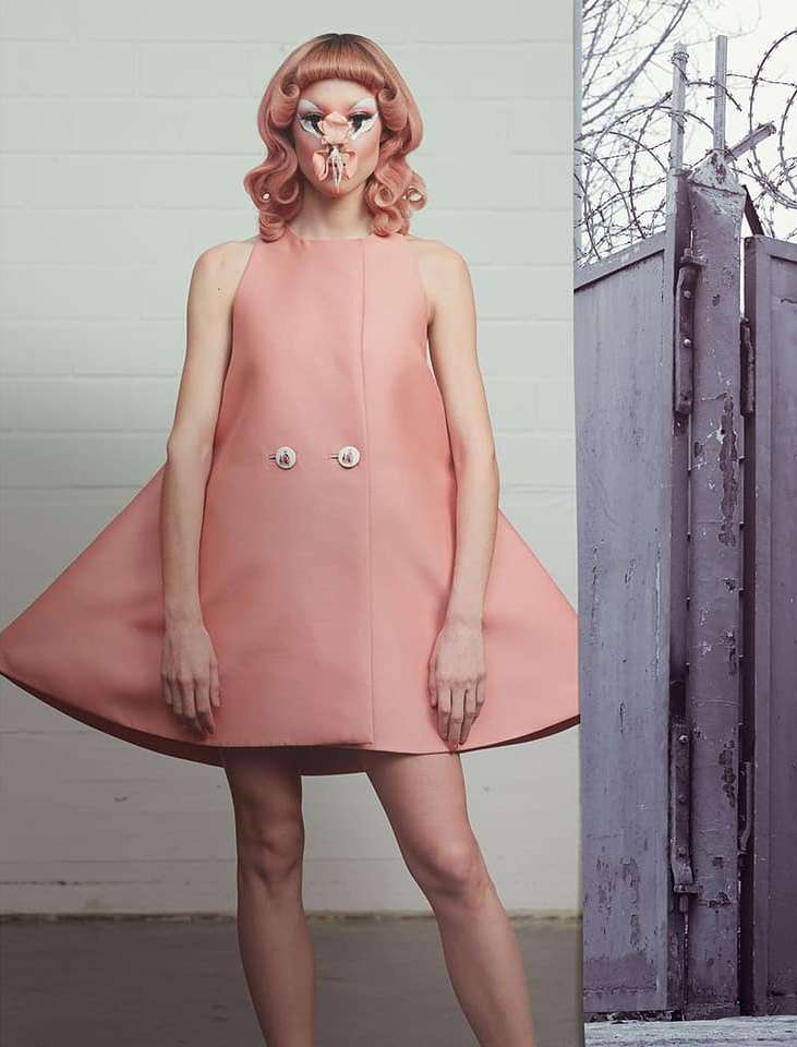 Fashion model with avant-garde makeup poses in a pink dress against a contrasting backdrop of a weathered metal gate and barbed wire.