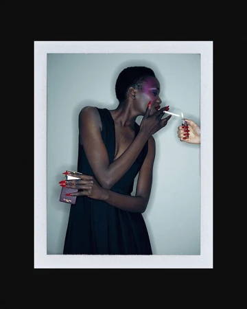 Elegant woman in a black dress lights a cigarette. Close-up fashion shot with vibrant pink eye makeup and red nails against a grey backdrop.