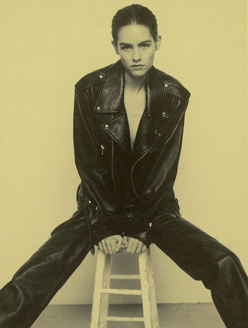 Model with slicked-back hair wearing a black leather jacket and trousers seated on a white wooden stool.