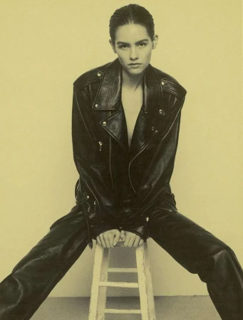 Model with slicked-back hair wearing a black leather jacket and trousers seated on a white wooden stool.