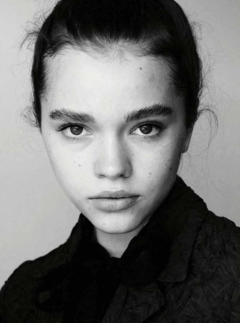 Monochrome close-up portrait of a young woman with dark hair and freckles wearing a dark textured jacket against a plain background.
