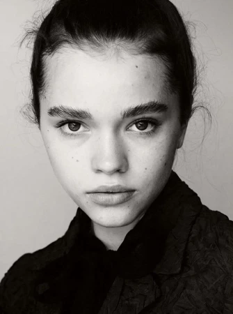 Monochrome close-up portrait of a young woman with dark hair and freckles wearing a dark textured jacket against a plain background.