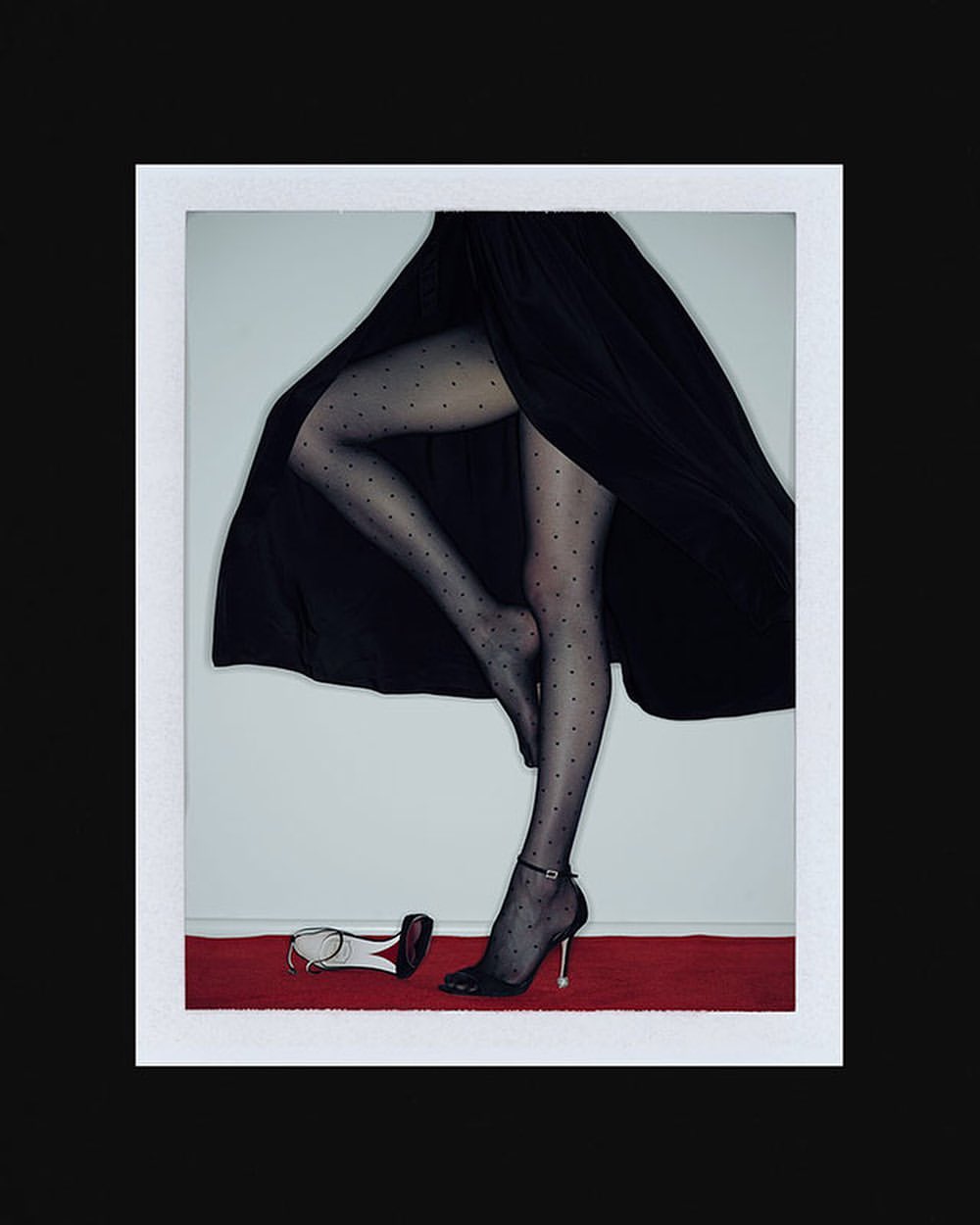 Elegant studio shot of a woman's legs in dotted stockings and black heels on a red carpet, with a matching shoe lying beside her.