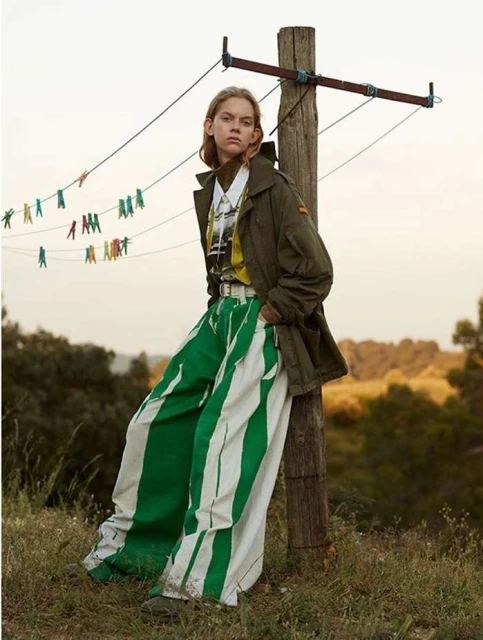 Young woman in striped pants leans against a weathered wooden post with a clothesline in a sunlit, grassy field with trees in the background.