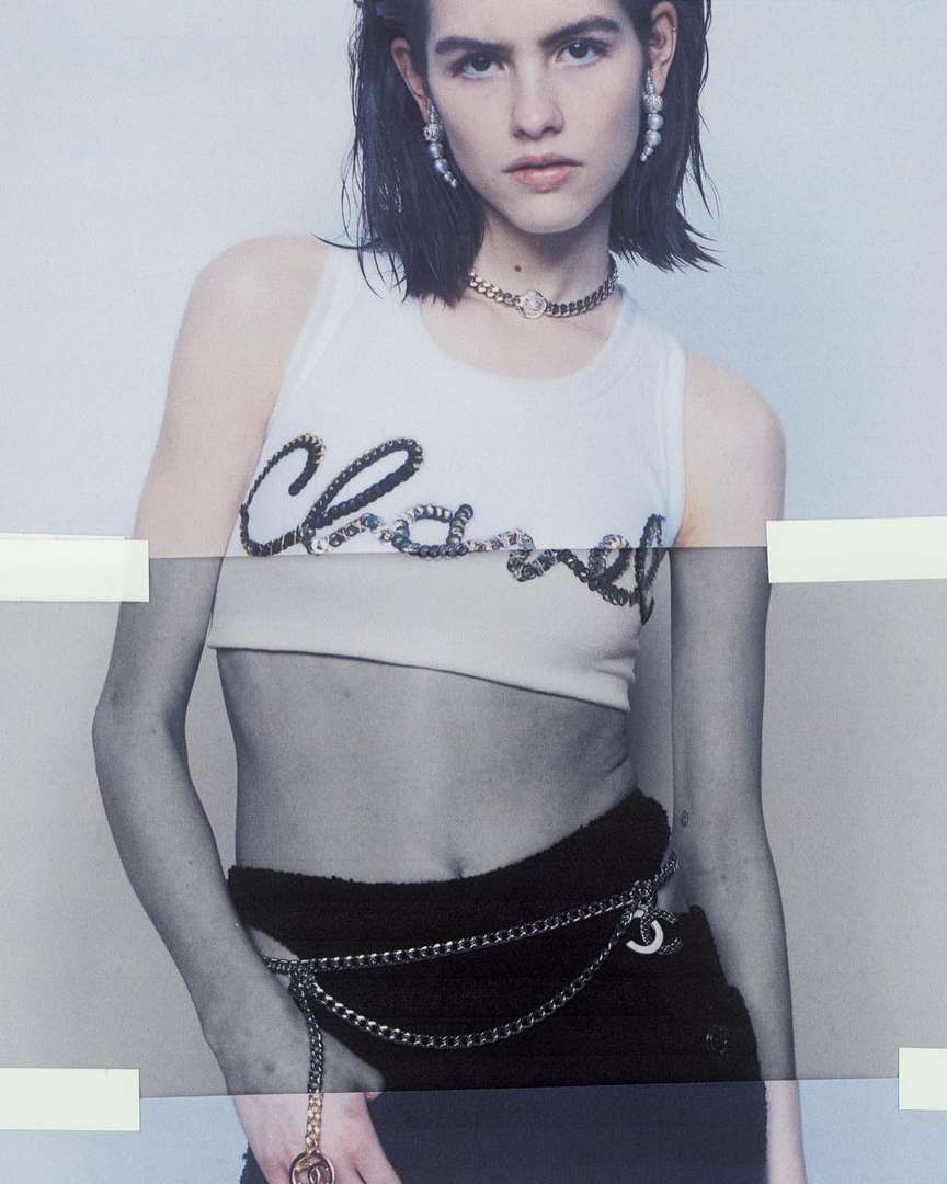 Female fashion model poses wearing a Chanel tank top and chain belt, accessorized with a necklace and pearl earrings in a studio shot.
