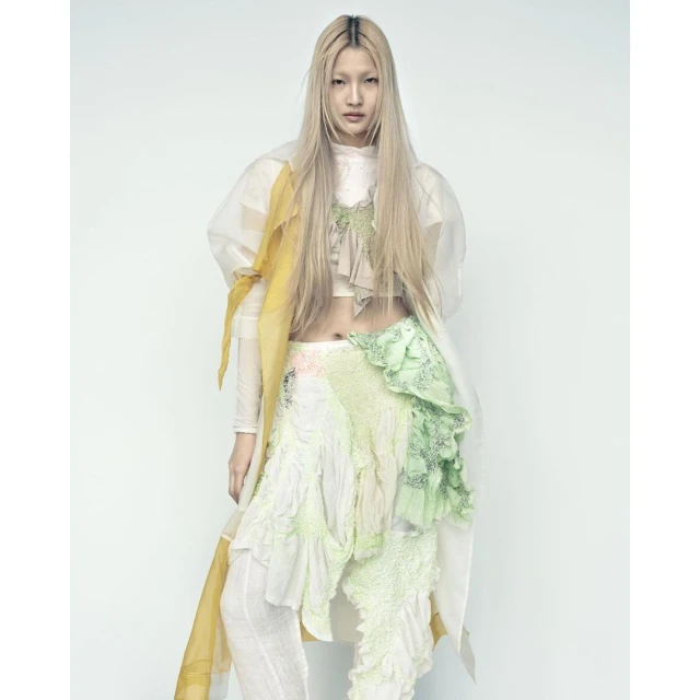 Woman with long blonde hair poses in a fashionable, ruffled, two-piece outfit in a studio with soft, diffused lighting. 