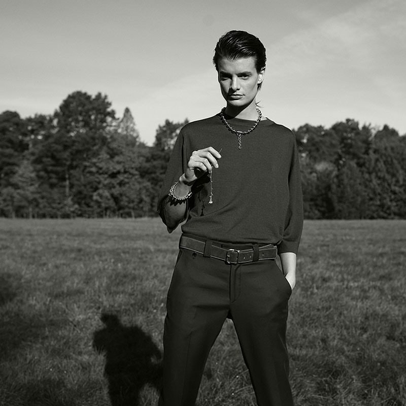 Monochrome portrait of a young adult wearing jewelry and holding a pendant necklace in a grassy field with trees in background.
