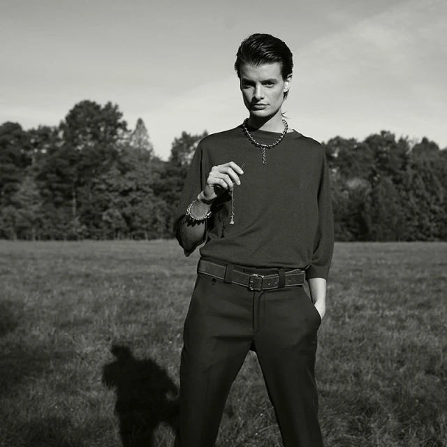 Monochrome portrait of a young adult wearing jewelry and holding a pendant necklace in a grassy field with trees in background.