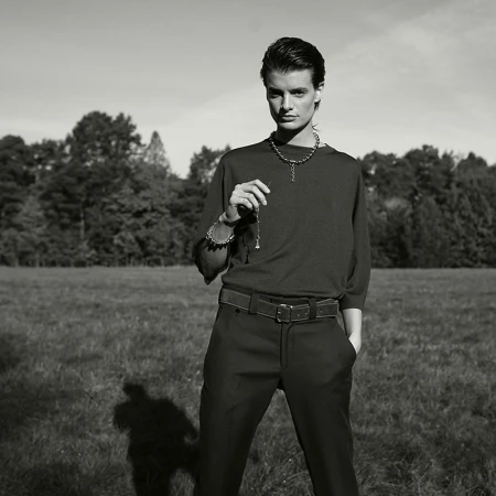 Monochrome portrait of a young adult wearing jewelry and holding a pendant necklace in a grassy field with trees in background.