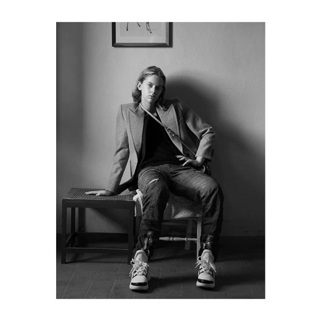 Black and white portrait of a young adult sitting on a bench, wearing a blazer, dark shirt, jeans and sneakers in an indoor setting.