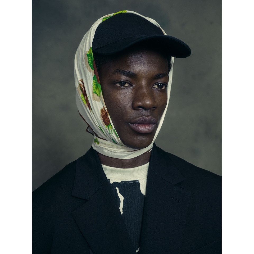 Portrait of an adult Black man in a black baseball cap and patterned headscarf, wearing a black blazer against a grey backdrop.